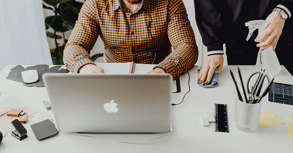 En kvinna torkar av ett skrivbord med rengöringsspray bredvid en person som arbetar vid en bärbar dator.