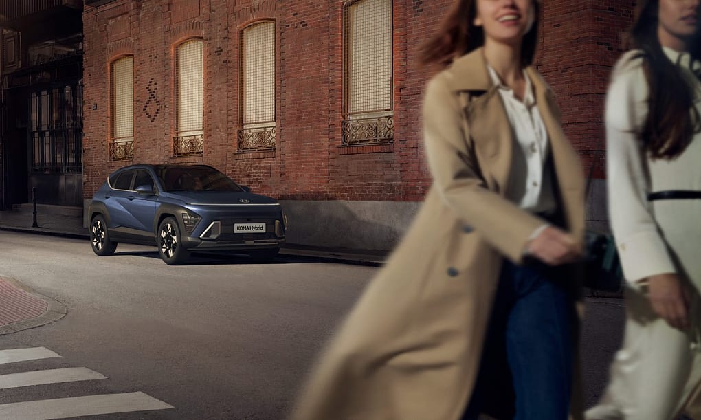 En blå Hyundai KONA Hybrid är parkerad på en stadsgata framför en tegelvägg med stora fönster. Två kvinnor, en i beige kappa och en i vit klänning, går förbi i förgrunden med glada uttryck. Bilden fångar en kvällsstämning med varm belysning och urbana detaljer.
