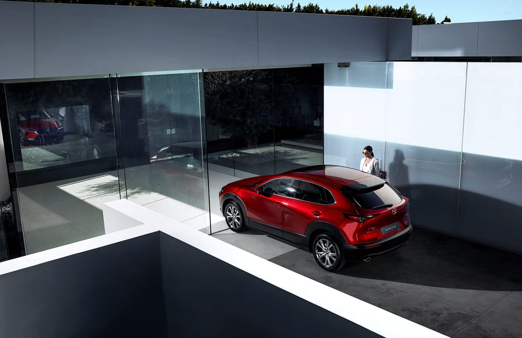 En röd Mazda står parkerad vid ett modernt hus med stora glaspartier. En kvinna står bredvid bilen.