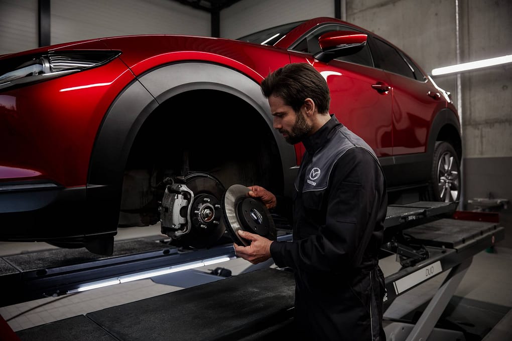 En bilmekaniker i Mazda-arbetskläder står i en verkstad och håller i en bromsskiva medan han arbetar på en röd SUV som är upphissad på en lyft. Fordonets framhjul är borttaget och verkstaden är belyst med kallt ljus i en industriell miljö med betongväggar.