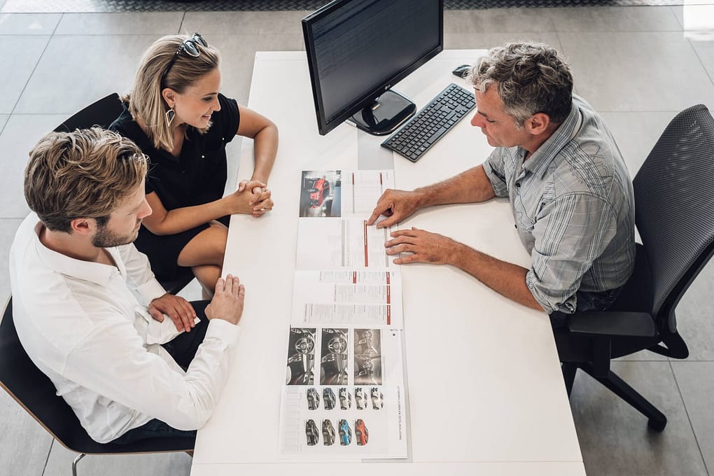 Ett par bläddrar i en bilkatalog hos en bilförsäljare. De sitter mittemot varandra och blir utbildade av försäljaren om modellerna de kan välja mellan.