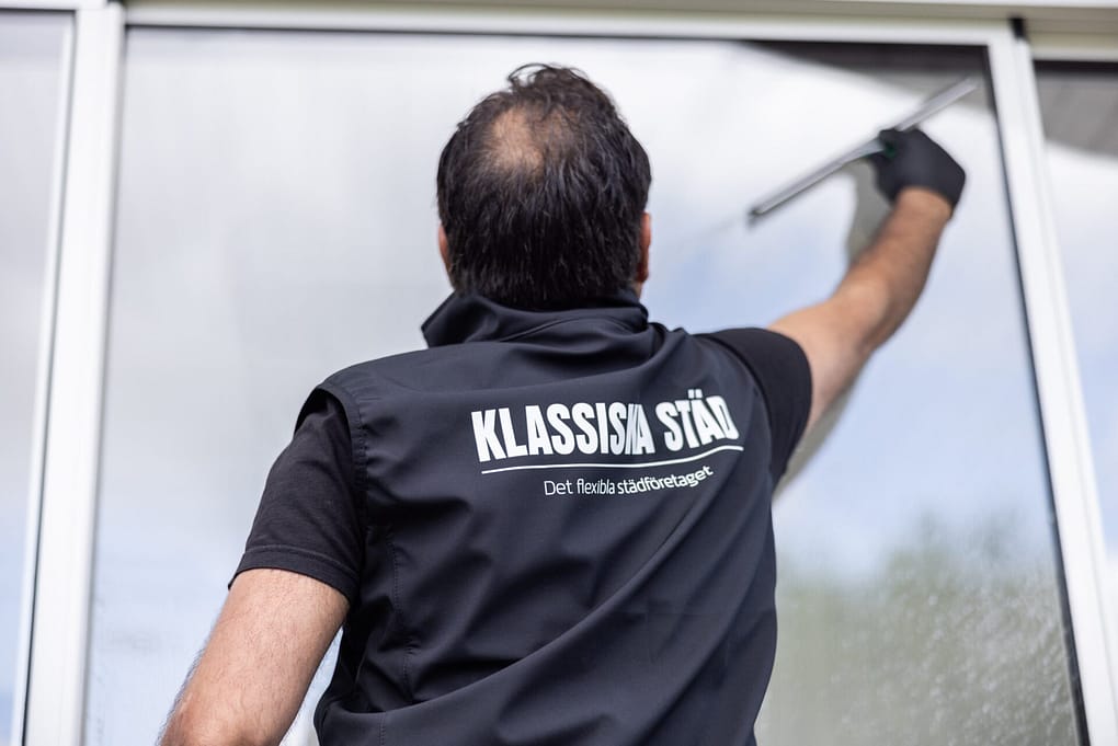 Medarbetare från Klassiska Städ putsar ett stort fönster utomhus, med logotypen synlig på ryggen av arbetsvästen.