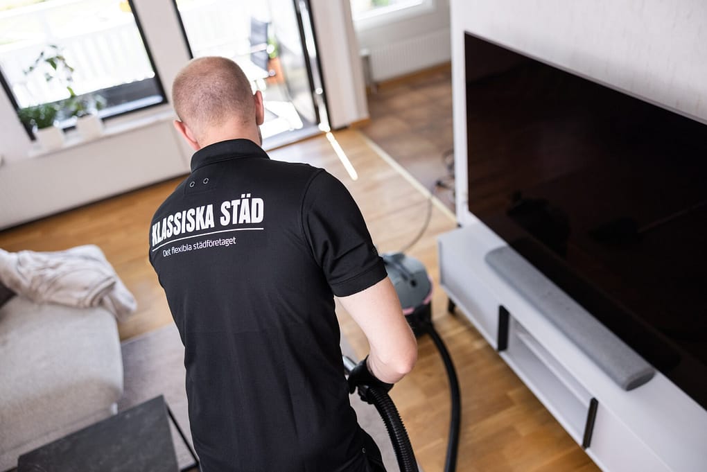 Städare från Klassiska Städ dammsuger ett vardagsrum framför en TV, med företagets logotyp synlig på ryggen av arbetskläderna.