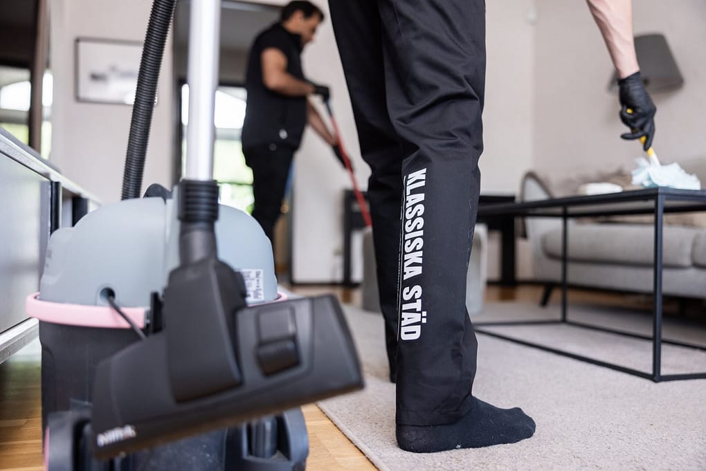 Städare från Klassiska Städ arbetar i ett vardagsrum, med fokus på dammsugare och företagslogotypen på byxbenet.