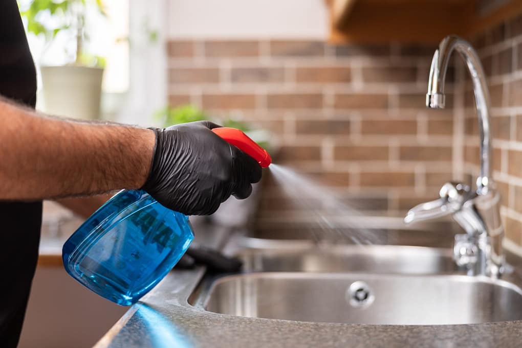 Städare från Klassiska Städ sprayar rengöringsmedel på en köksbänk intill en diskho, iförd svarta skyddshandskar.
