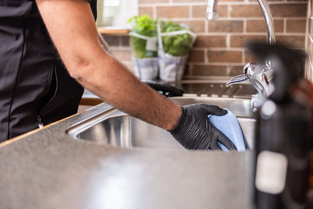 Städare från Klassiska Städ rengör en diskho i ett kök med en blå mikrofiberduk, iförd svarta skyddshandskar.