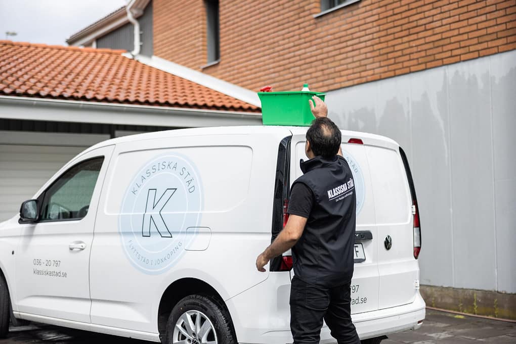 Medarbetare från Klassiska Städ placerar en grön städlåda på taket av företagets vita servicebil utanför en byggnad.