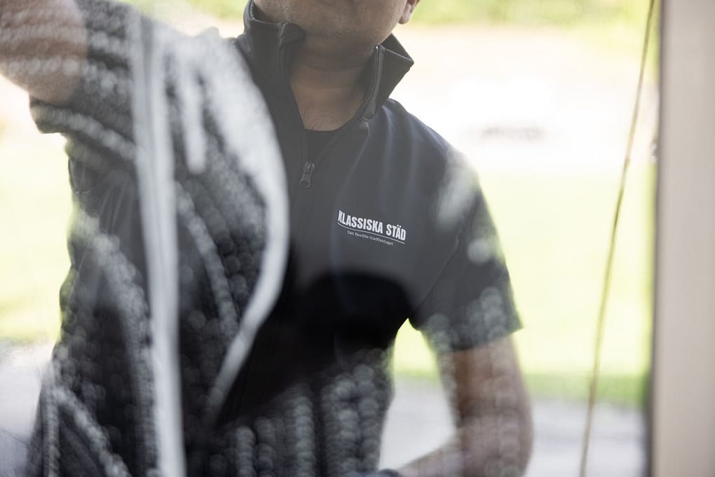 Närbild genom glaset på en städare från Klassiska Städ som putsar ett fönster med rengöringsmedel.