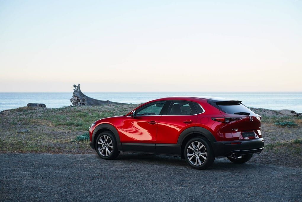 En röd Mazda CX-30 är parkerad vid en kust med utsikt över havet. Bilen står på en grusväg med gräs och drivved i bakgrunden. Himlen är ljusblå och solen håller på att gå ner, vilket skapar ett lugnt och naturnära stämningsläge.
