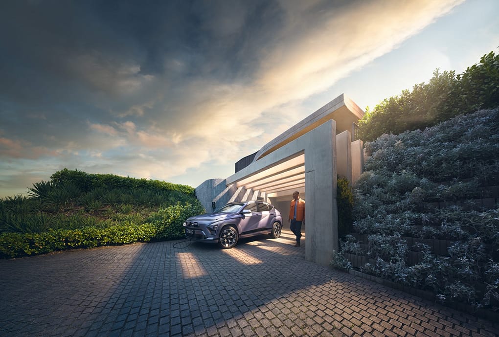 En modern villa med en uppfart av stenplattor där en blå SUV står parkerad under ett betongtak. Bilen laddas vid en elstation, och en man i orange jacka kliver ut från garaget. Omgivningen är grönskande och kvällssolen lyser genom molnen och skapar en varm stämning.