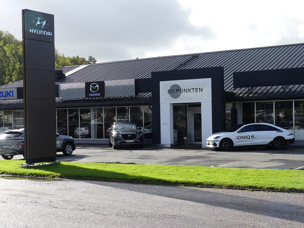 En bilhall med skyltar för Hyundai, Mazda och Suzuki ses från parkeringen utanför. På byggnaden står det "BILPUNKTEN" ovanför entrén. Flera bilar står parkerade framför, bland annat en vit Hyundai IONIQ 6 med texten "IONIQ 6." tryckt på sidan. Grönt gräs och våt asfalt syns i förgrunden under en mulen himmel.