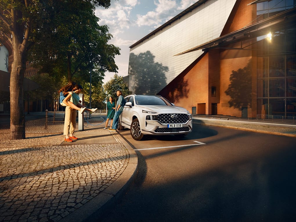 En vit Hyundai SUV står parkerad vid en trottoar i en stadsmiljö med moderna byggnader. En familj med två vuxna och två barn står vid bilen, som är ansluten till en laddstation. Solen skiner mellan träden och skapar en varm och livlig känsla.