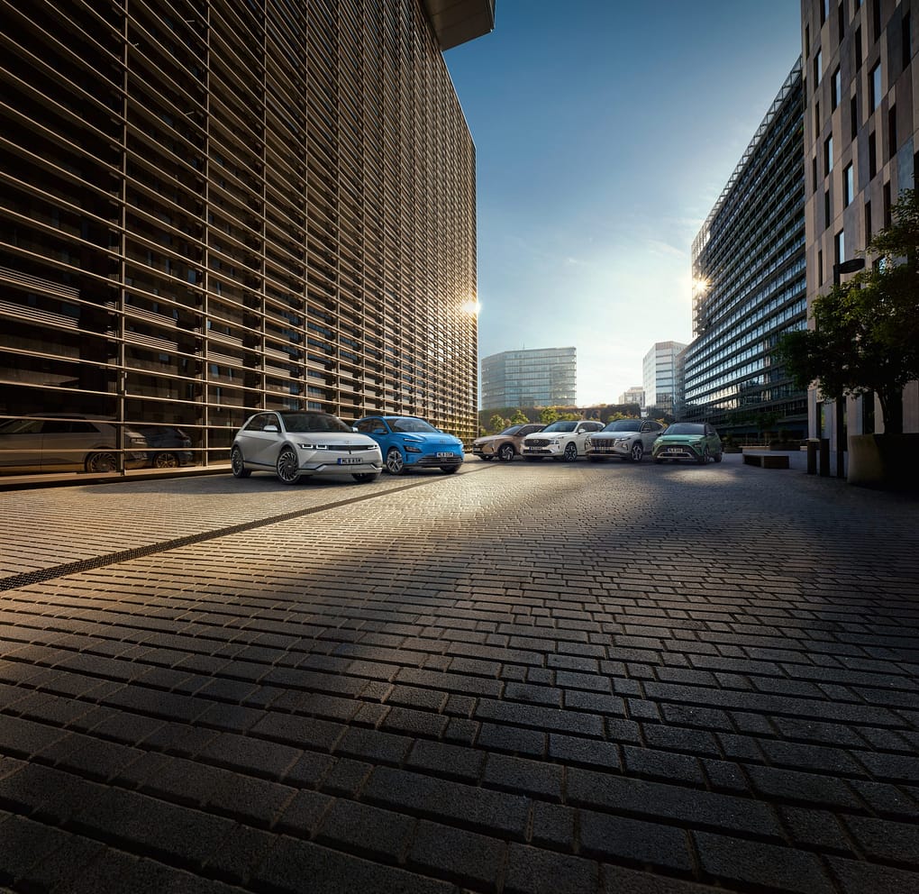 En rad moderna bilar från Hyundai står parkerade på en stenlagd gata mellan höga kontorsbyggnader med glas- och metallfasader. Morgonsolen lyser in från horisonten och reflekteras i byggnadernas fönster, vilket skapar en varm stadsatmosfär.