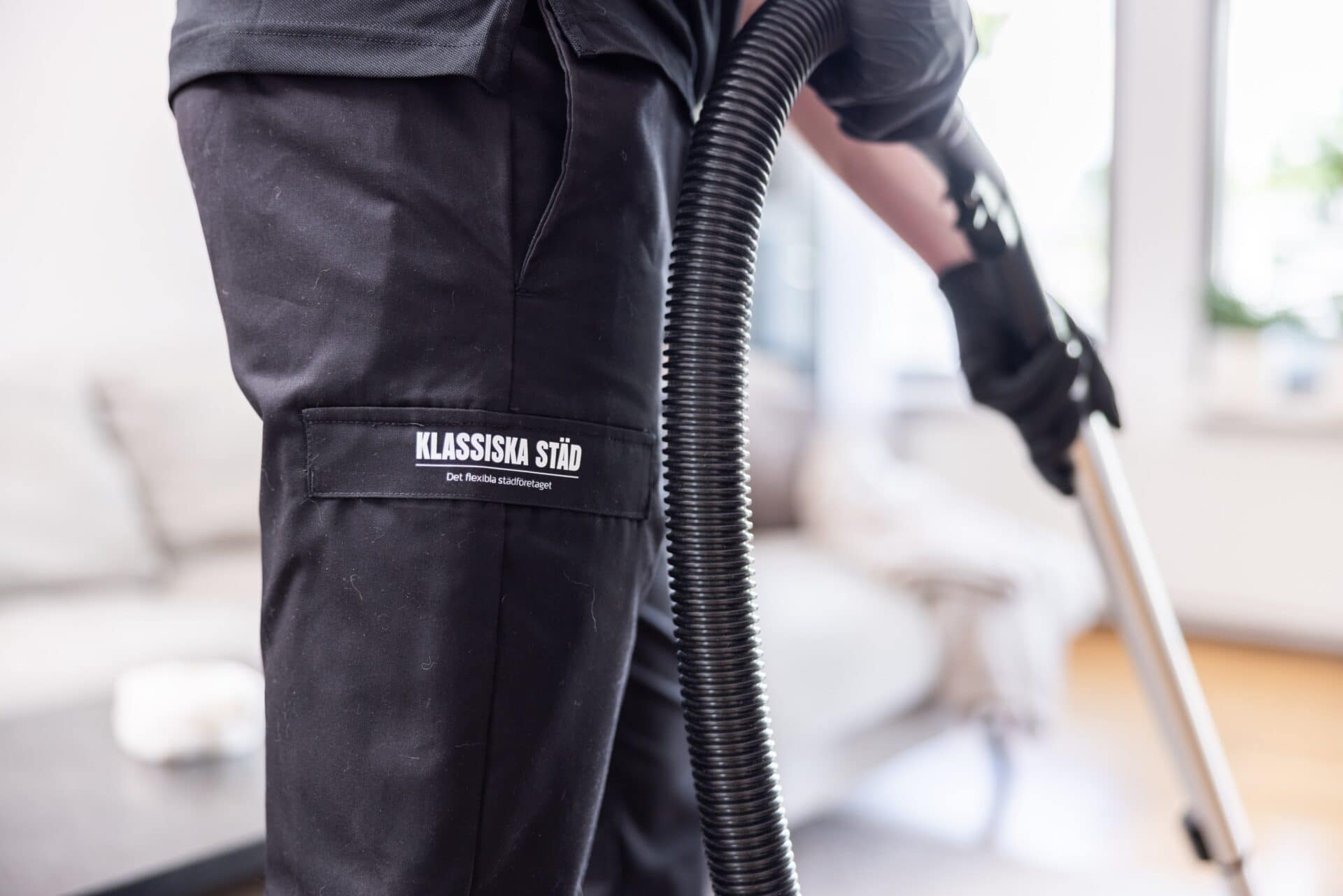 Underdelen av en städare som dammsuger en bostad. Företagslogotyp syns på byxan.