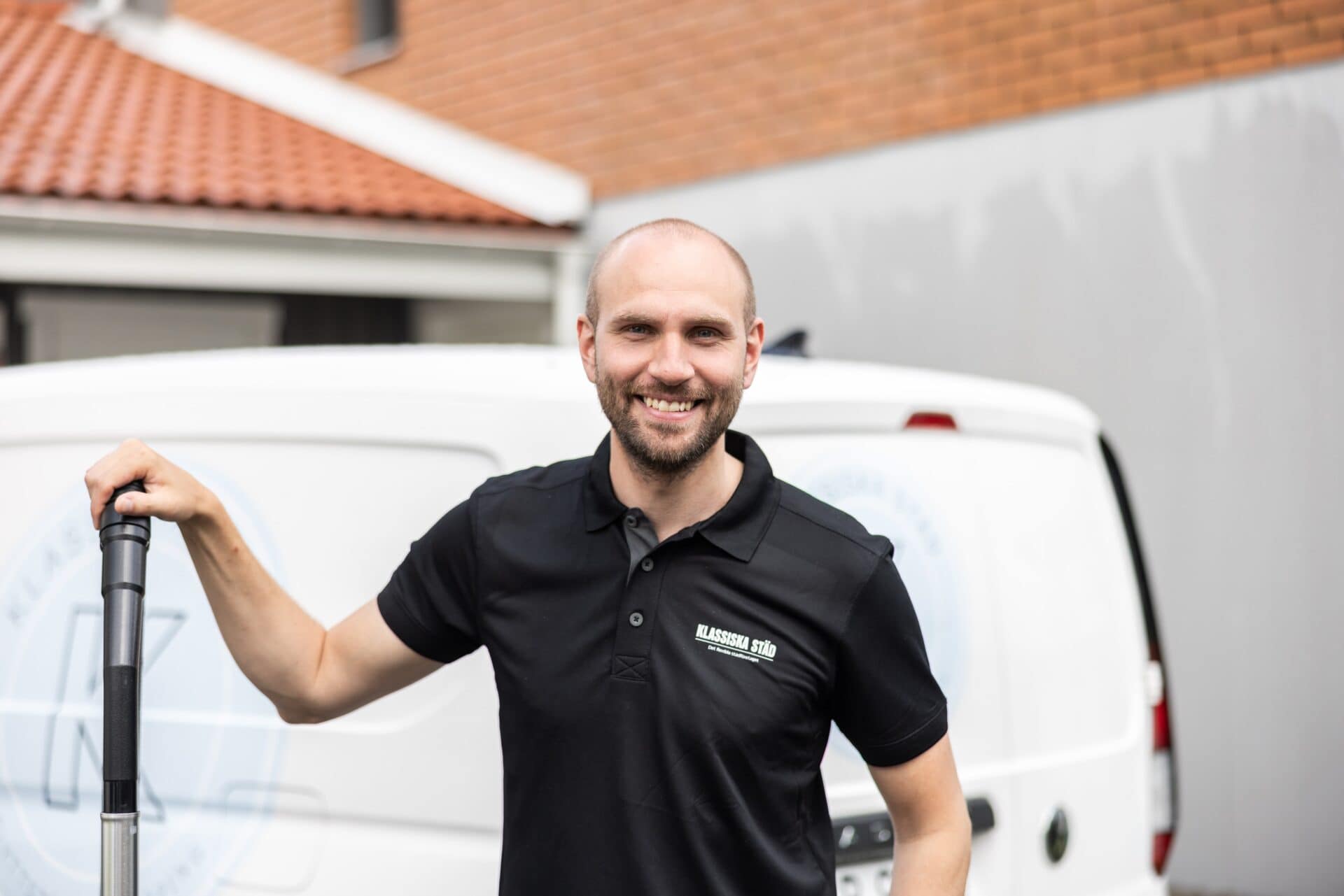 Leende städare från Klassiska Städ står framför en vit firmabil, iförd svart pikétröja med företagets logotyp.