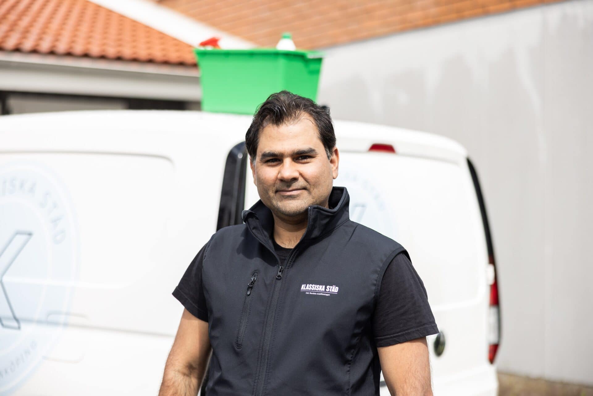 Medarbetare från Klassiska Städ står framför en vit firmabil med en grön städlåda på taket, iklädd svart arbetsväst med logotyp.