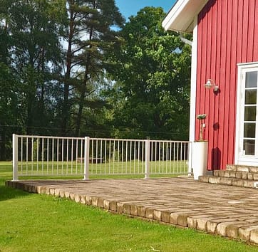 Vitt aluminiumräcke vid uteplats till ett rött hus med vita knutar.