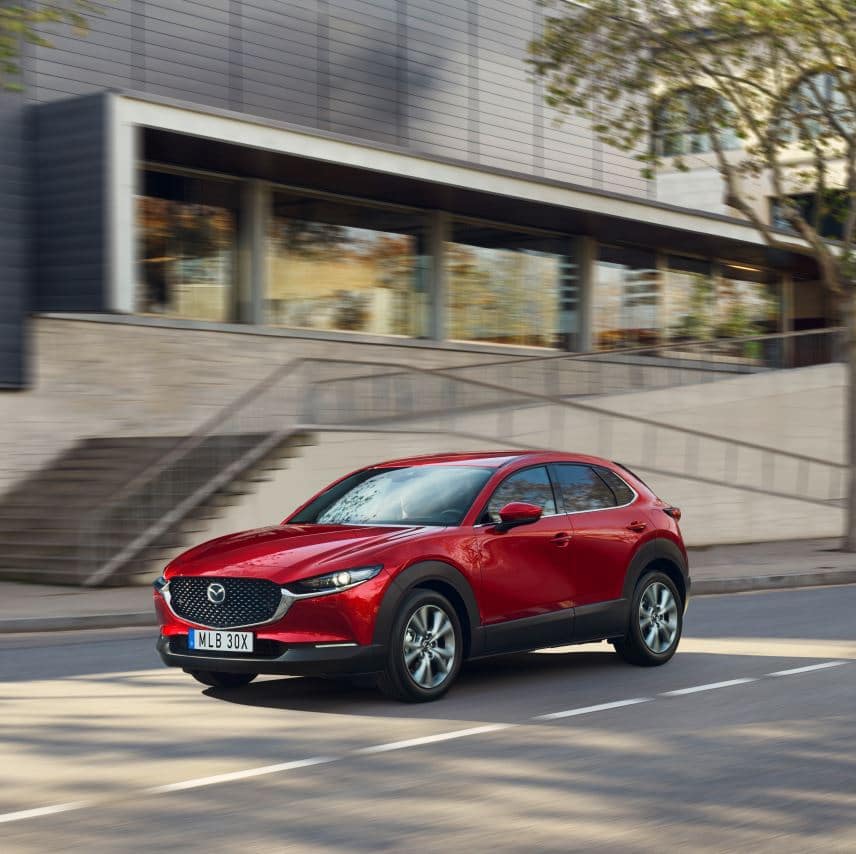 En röd Mazda CX-30 kör på en stadsgata framför en modern byggnad med stora fönster och trappor. Bilen har ett elegant och sportigt utseende och rör sig i dagsljus under trädens skuggor.