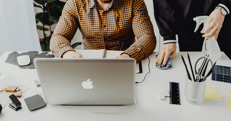 En kvinna torkar av ett skrivbord med rengöringsspray bredvid en person som arbetar vid en bärbar dator.