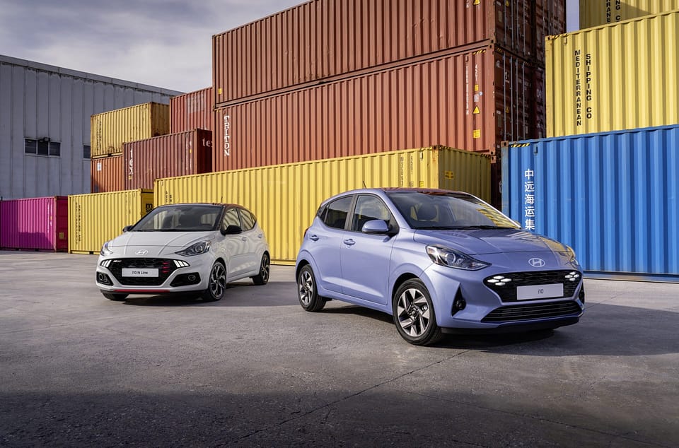 Två Hyundai i10-bilar står parkerade framför färgglada containrar. Den ena är ljusblå med enkel och rundad design, medan den andra är vit med sportiga svarta detaljer och röda inslag. Platsen ser ut som ett industriområde med staplade containrar i bakgrunden.