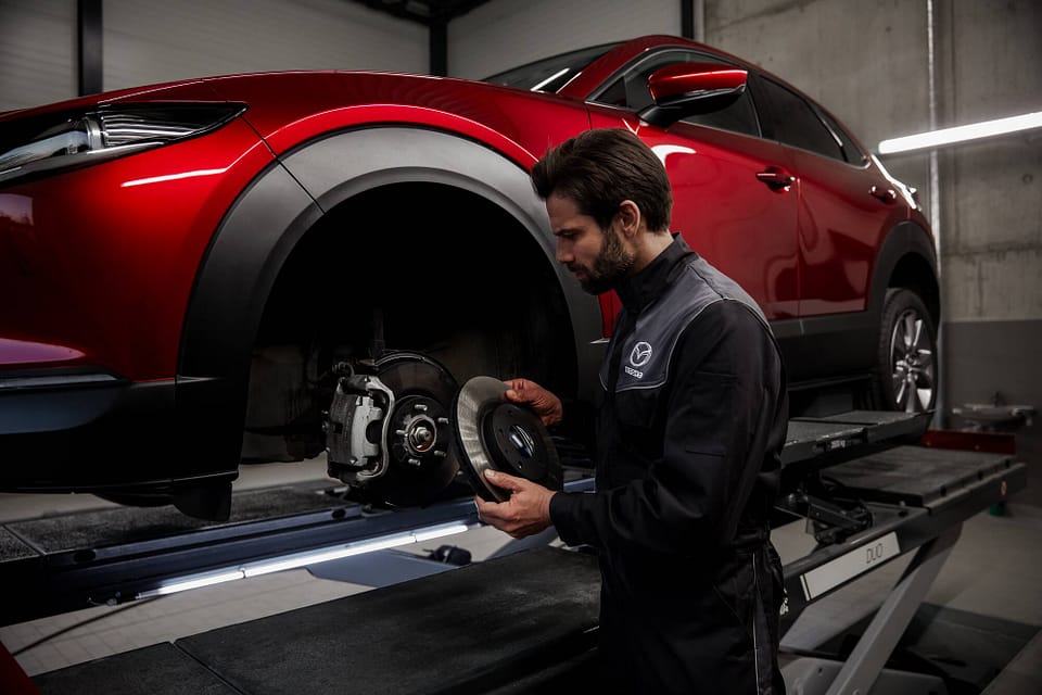 En bilmekaniker i Mazda-arbetskläder står i en verkstad och håller i en bromsskiva medan han arbetar på en röd SUV som är upphissad på en lyft. Fordonets framhjul är borttaget och verkstaden är belyst med kallt ljus i en industriell miljö med betongväggar.