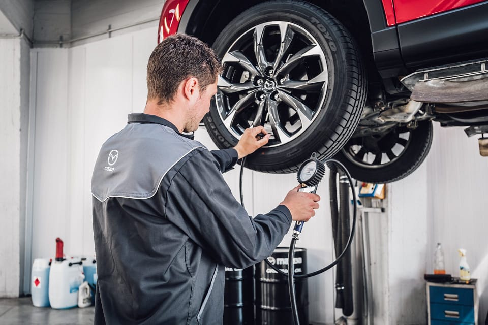 En bilmekaniker i Mazda-arbetskläder kontrollerar lufttrycket i ett däck på en röd bil som är upphissad i en verkstad. Han använder en tryckmätare med slang och står fokuserad framför det blanka fälghjulet. Verktyg och oljefat syns i bakgrunden.