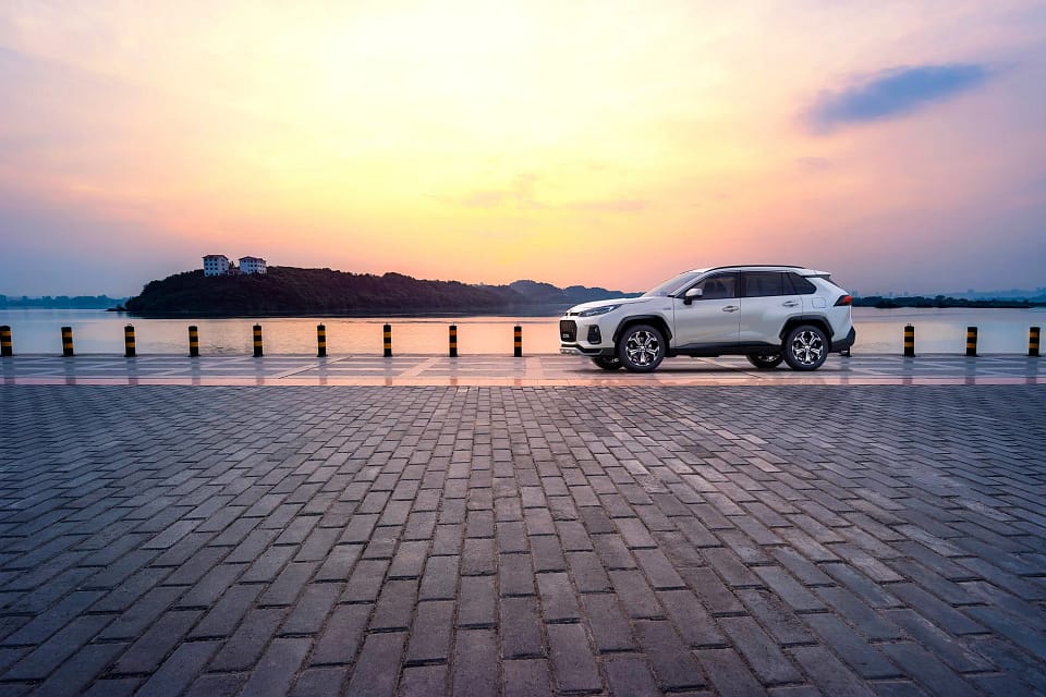 En vit bil sett från sidan står parkerad vid en hamn i solnedgången.