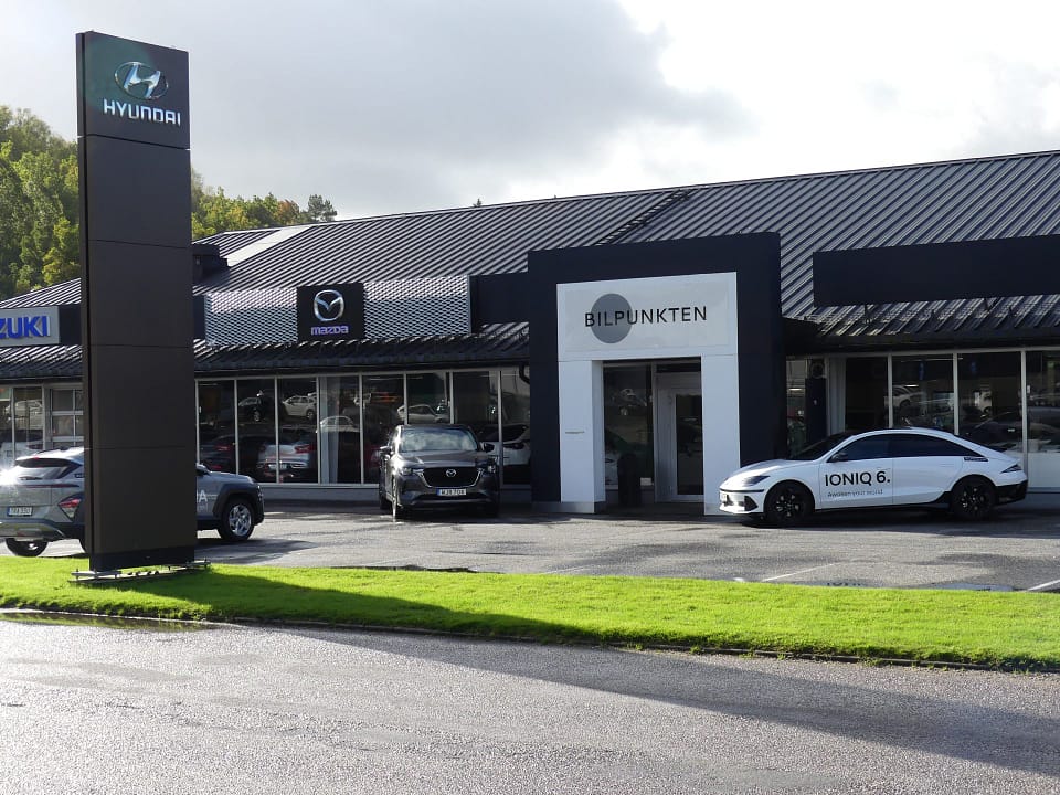 En bilhall med skyltar för Hyundai, Mazda och Suzuki ses från parkeringen utanför. På byggnaden står det "BILPUNKTEN" ovanför entrén. Flera bilar står parkerade framför, bland annat en vit Hyundai IONIQ 6 med texten "IONIQ 6." tryckt på sidan. Grönt gräs och våt asfalt syns i förgrunden under en mulen himmel.