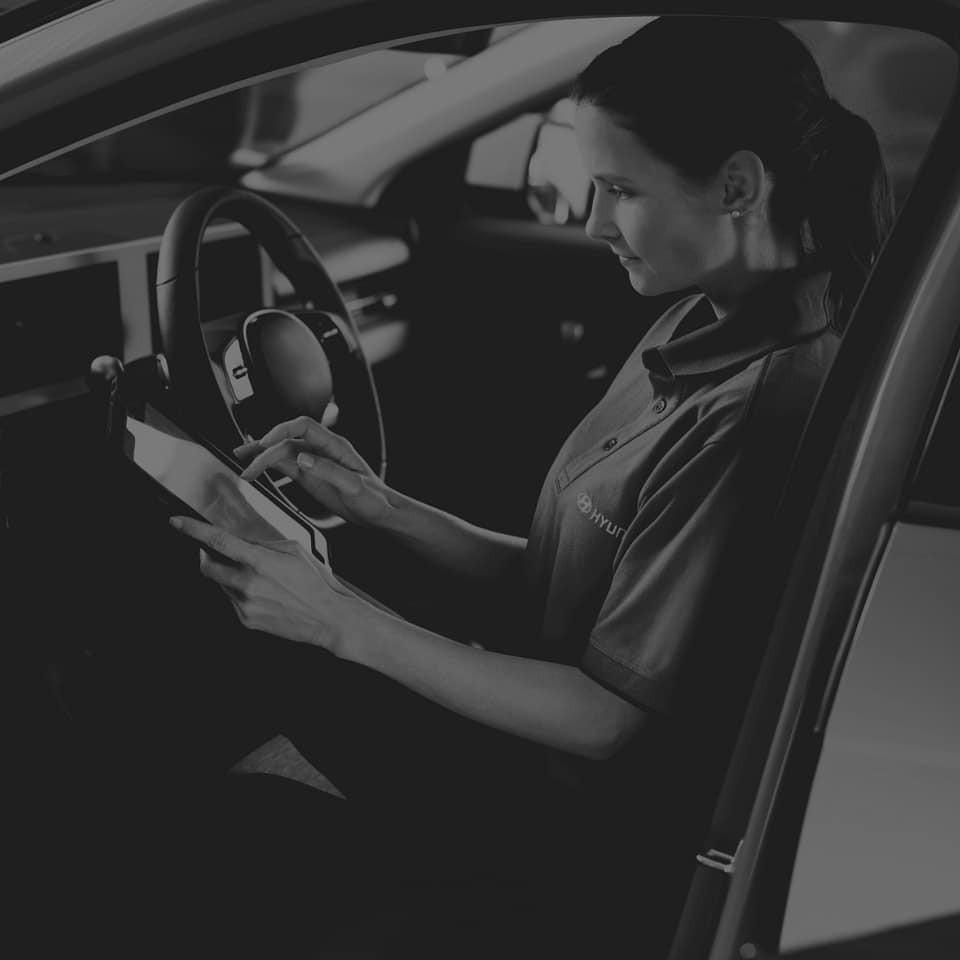 En kvinna i Hyundai-arbetskläder sitter i förarsätet på en bil och använder en surfplatta. Hon ser fokuserad ut och verkar utföra en digital kontroll eller diagnos. Bilden är i svartvitt och har en professionell ton.
